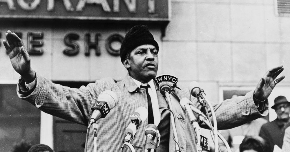 Black and white image of Bayard Rustin speaking at a podium with his arms out wide.
