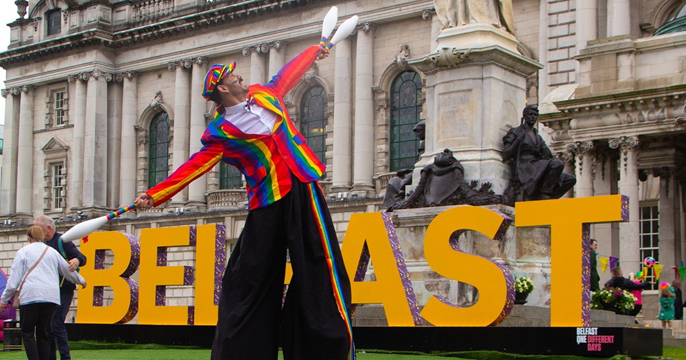 An image from Belfast Pride family fun day of a person in stilts and a rainbow jacket and hat in front of a Belfast sign.