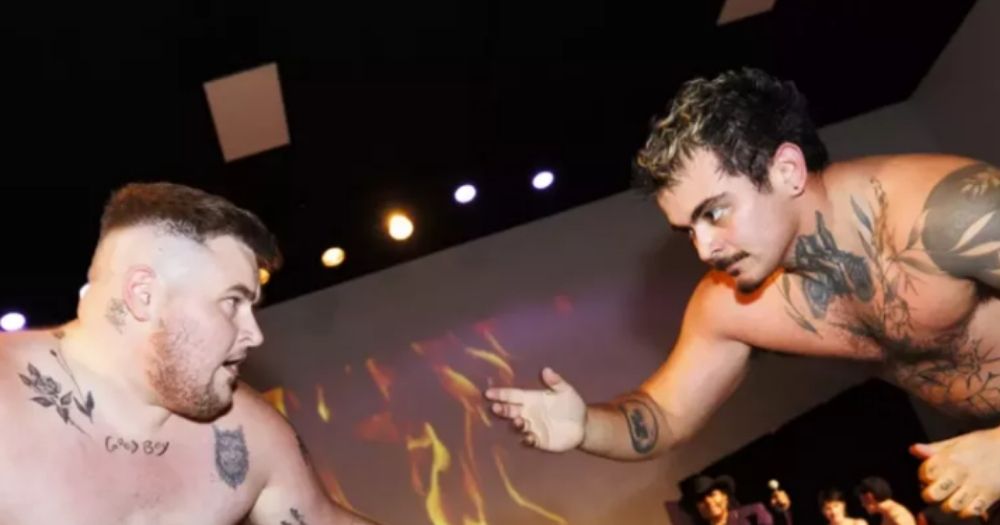 T boy wrestling competitors, prepare to hook in a fighting stance, in front of a screen showing flame, there is a gentleman in the background holding a musical device like a guitar or violin.