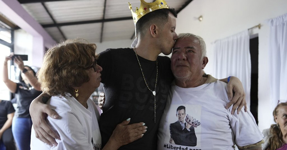 Gay man Andry Hernández Romero reunited with his parents after being freed from an El Salvador prison. He wears a black t-shirt and a gold crown and kisses his father on the head. He has his other arm around his mother.
