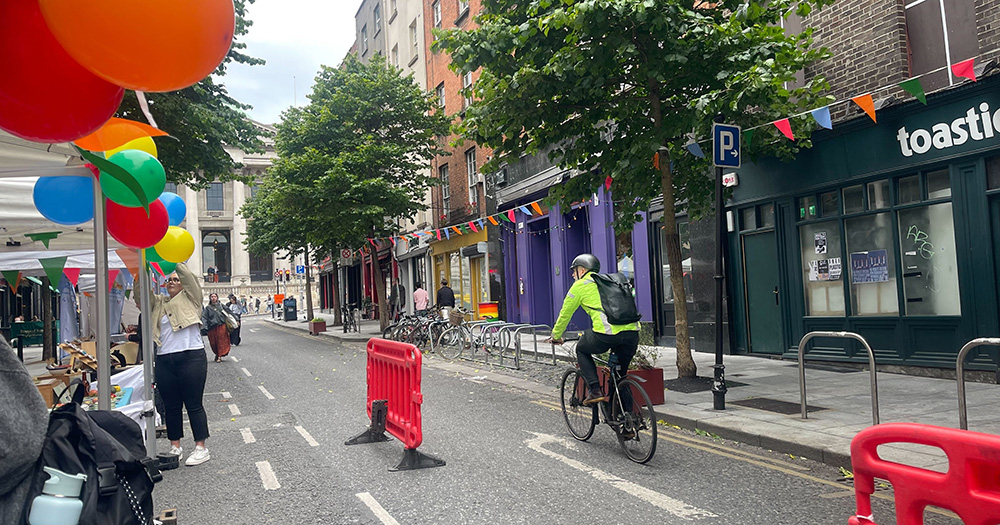 An image of traffic-free Parliament Street. There are market stalls with colourful balloons attached to them, and a cyclist cycling down the street.