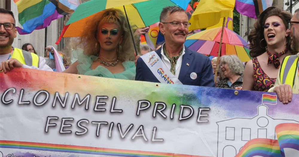 Marchers at Clonmel Pride in 2024. They hold a banner reading Clonmel Pride Festival, and march with rainbow flags and umbrellas held over them.
