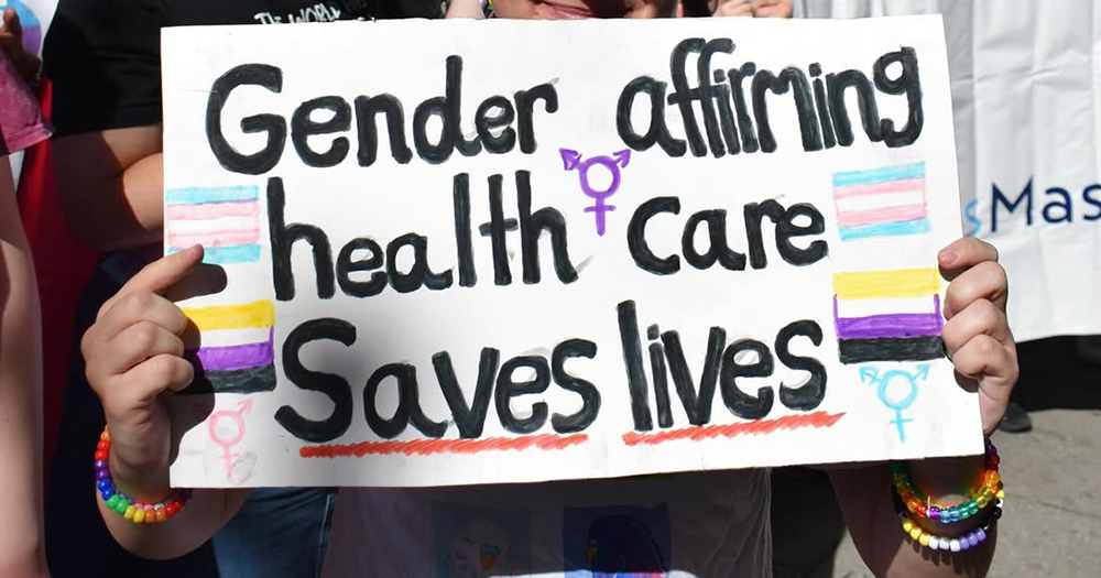 This article is about INMO advocating for inclusive trans healthcare. In the photo, a person holding a sign that reads "Gender affirming health care saves lives".
