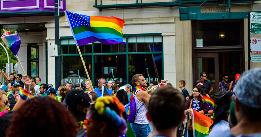 This article is about the Trump administration restoring funding to LGBTQ+ and HIV organisations. The image shows a Pride parade, with a rainbow and US flag combined.