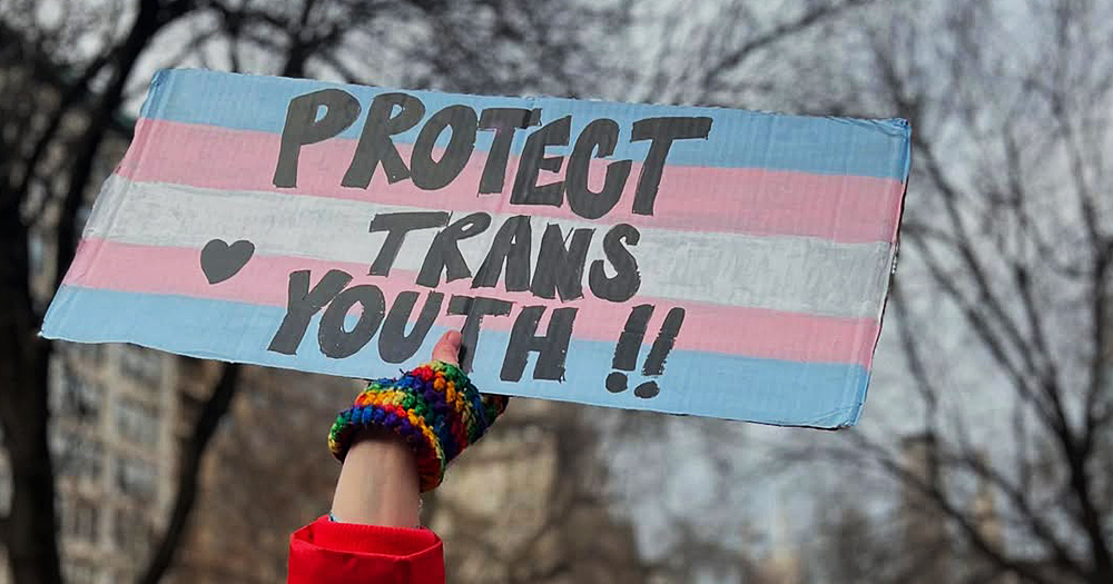 This article is about the closure of a US gender-affirming care centre for trans youth. In the photo, a hand holding up a sign that reads "Protect Trans Youth".
