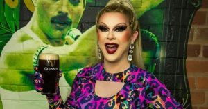 Drag queen Bonnie Ann Clyde wearing a purple leopard print shirt and holding a Guinness, in front of a Freddie Mercury mural.