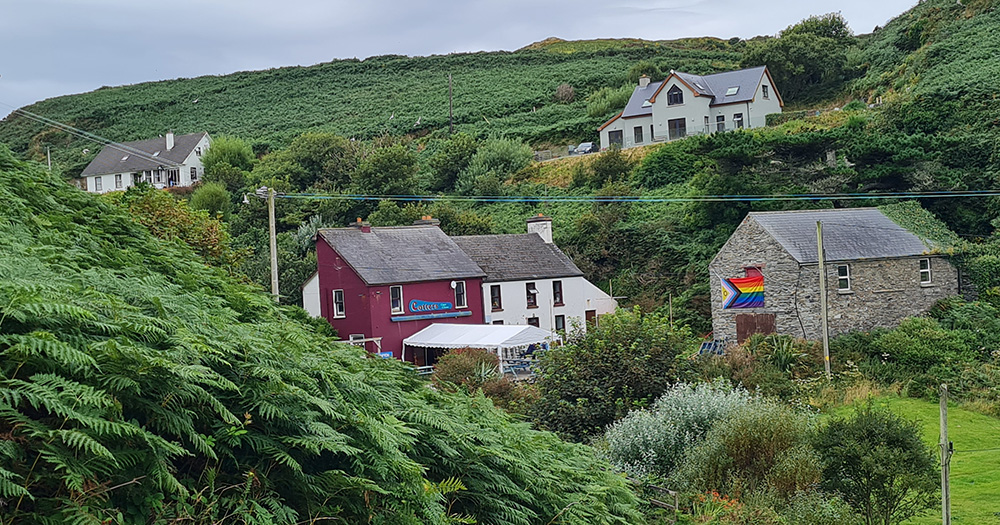 An image from Bród Chléire. It shows green hills and fields, with some houses - one with an LGBTQ+ flag.