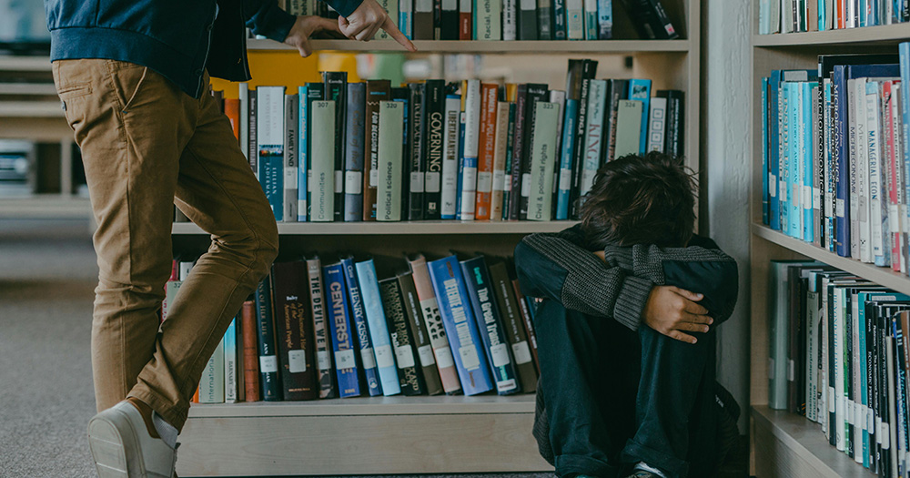 A child facing bullying. He sits on the floor of a library with his head in his arms as someone stands beside him pointing at him.