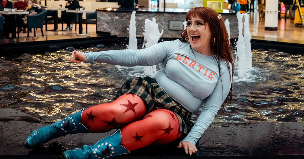 Irish singer CMAT, who has just announced her biggest show ever, leaning on a fountain and smiling, while wearing bright red stockings with big stars and blue cowboy boots.
