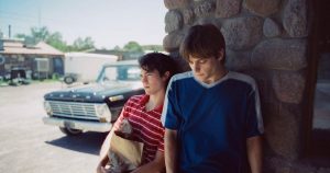 The image shows Conan Gray and Corey Fogelmanis, from the Caramel music video on the album Wishbone. The pair are standing next to each other against the background of a brick wall. An old car can be seen in the background. Corey is looking at the floor and wearing a blue t-shirt. Conan is staring off-camera, wearing a red and white striped shirt and holding a brown paper bag of groceries.