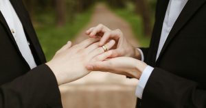 A drag queen has been officiating queer weddings in the US. The image shows two people in suits, one putting a ring on the other's finger.