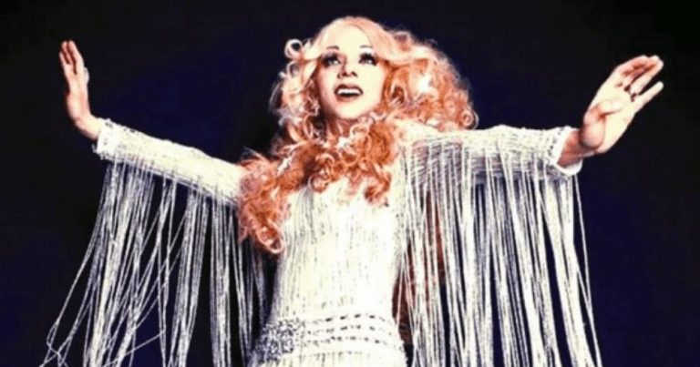 This image shows the Lebanese drag queen Bassem Feghali in a sparkly silver dress with her arms outstretched. She has blonde curly hair and bangs and is performing to an audience.