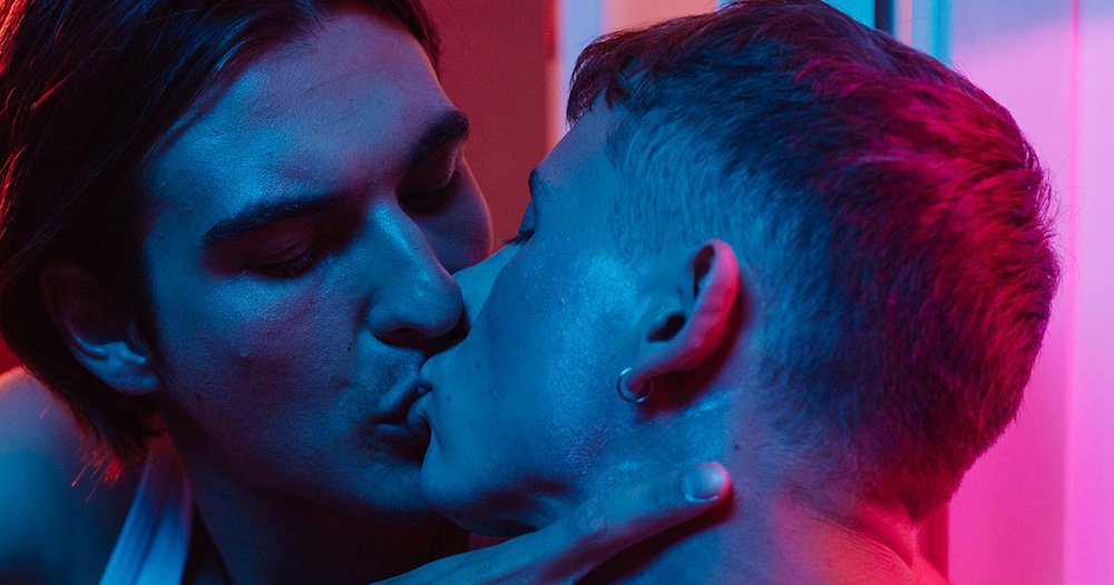 This article is about the new Grindr hub of original content. The image shows a close up of two men kissing under blue lights.
