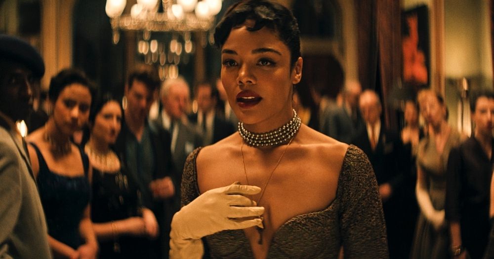 Actor Tessa Thompson starring in the film Hedda, wearing an elegant 1950s dress in a room full of people at a party.