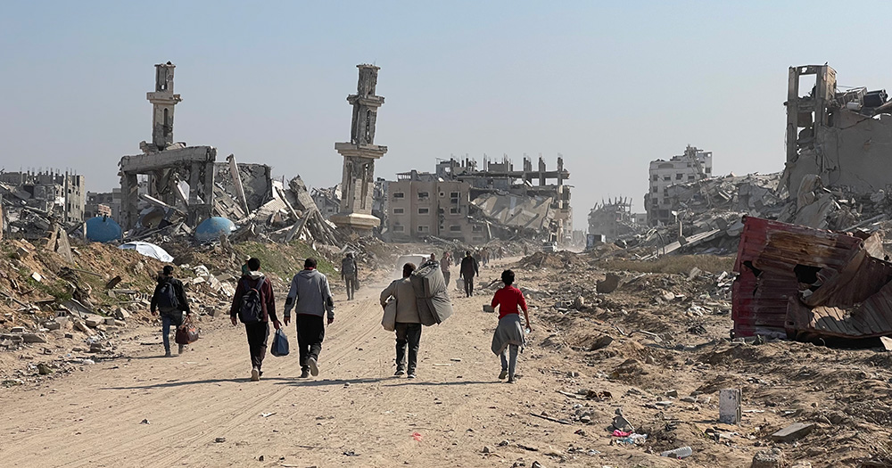 People walking through Gaza, which Israel has destroyed. Destroyed buildings are visible in the background.