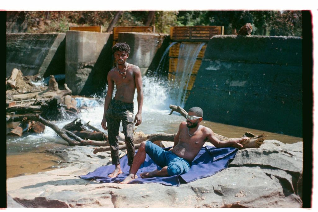Two young black people pose shirtless on a sunny day on front of a manmade waterfall. One lays on a blanket while the other stands and looks at the lens of the camera.