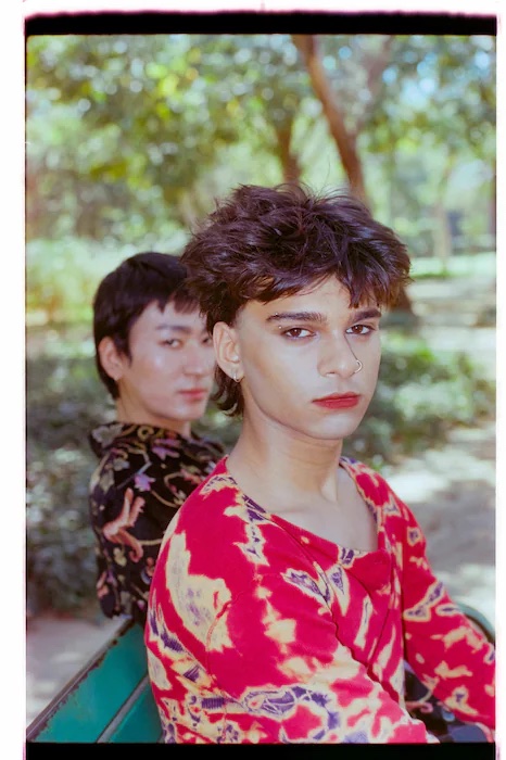 Two young people in feminine attire and make up look at the camera. They appear to be sitting in a park.