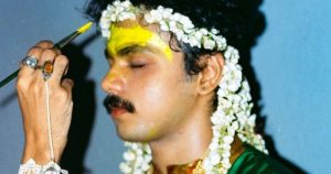 A portrait of an Indian youth wearing a flower headdress. A hand reaches out and paints their forehead yellow. They are at a queer, trans, and non-binary event.