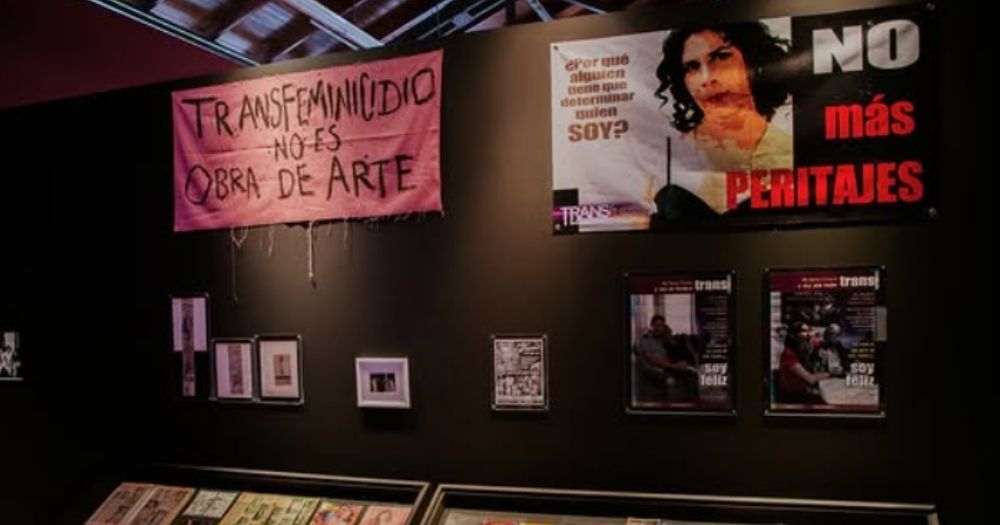 The image shows an exhibition of transfeminine art. There are two display cabinets filled with objects as well as several posters and framed photos hanging on a black wall.