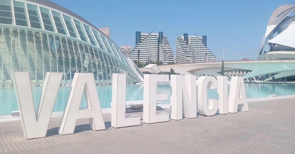 A white Valencia sign in front of the Ciudad de las Artes y las Ciencias. In the background you see a glass building and pond. Valencia will host the Gay Games in 2026.