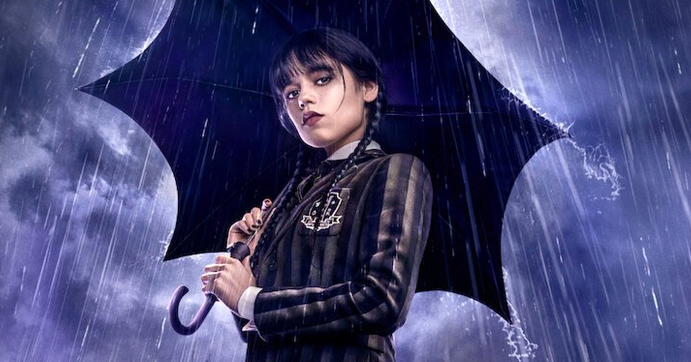 Main character of Wednesday tv show, holding a black umbrella with a rainy background.
