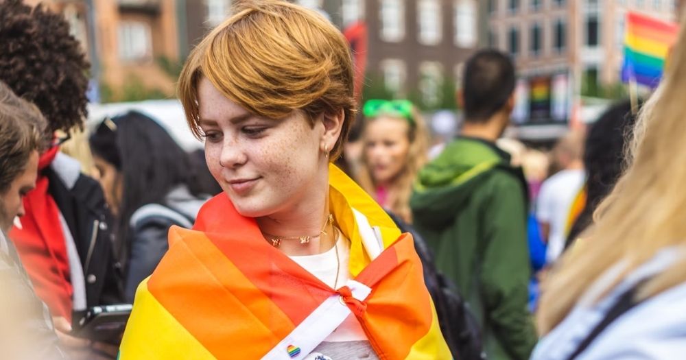 This article is about a new mental health hub for LGBTQ+ youth. In the photo, a young person smiling softly while looking down and wearing a rainbow flag on their shoulders.