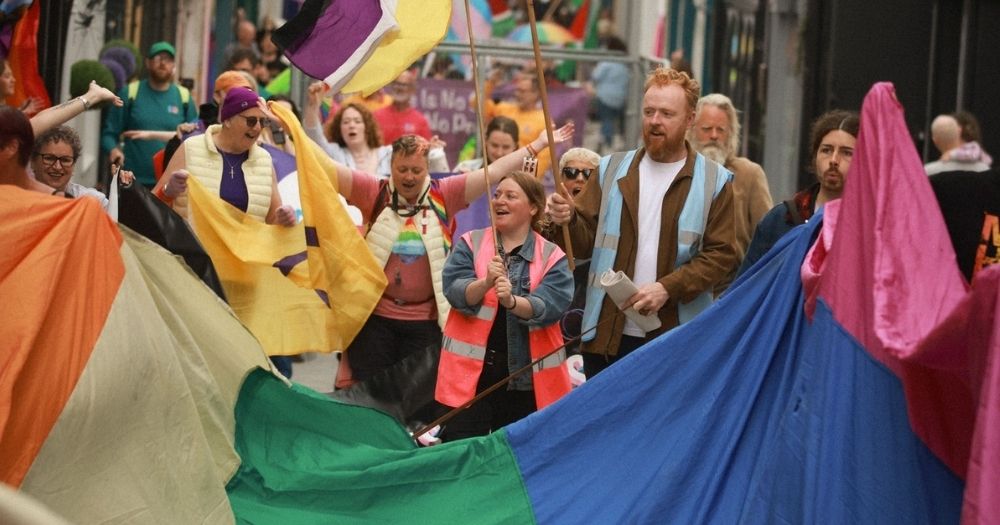 Image from Clare Pride march 2024. People hold a big rainbow flag while stewards in high-vis vests cheer and wave flags.