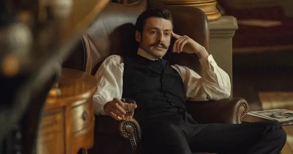 The image shows Arthur Guinness from the TV show House of Guinness. He is pictured sitting in a large armchair, with a glass in his hand. He is wearing a black suit, waistcoat, and white shirt, and has a mustache. He is holding his right hand up to his temple.