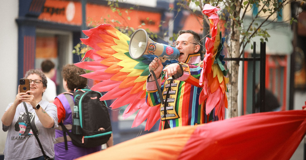 An image of Eddie McGuinness, one of the organisers of the West of Ireland's EuroPride 2028 bid. He speaks into a megaphone, wearing a rainbow suit and rainbow wings.