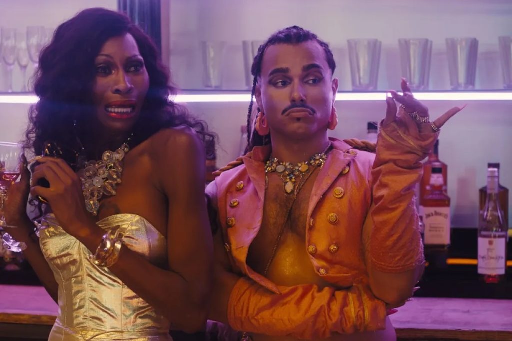 A drag queen and fashionable person stand at a bar. The queen looks frightened in a gold dress, the other person looks non-chalant in an incredible orange biker style jacket. A still from Queens of the Dead. Queer halloween costume ideas 2025.