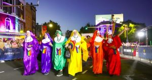 This image shows the Sydney order of the Sisters of Perpetual Indulgence taking part in a parade. There are 6 nuns dressed in in colourful habits and holding pride flags, walking along the road with crowds gathered on either side.