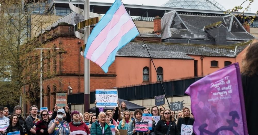 This article is about Trans Pride NI in Belfast. In the photo, people gathered for a rally in Belfast holding signs with protest slogans and trans flags.