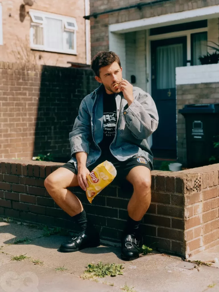 Jonathan Bailey sits on a short brick wall outside a two-up two-down type of house, as he wears short shorts, a denim jacket, and eats crisps.