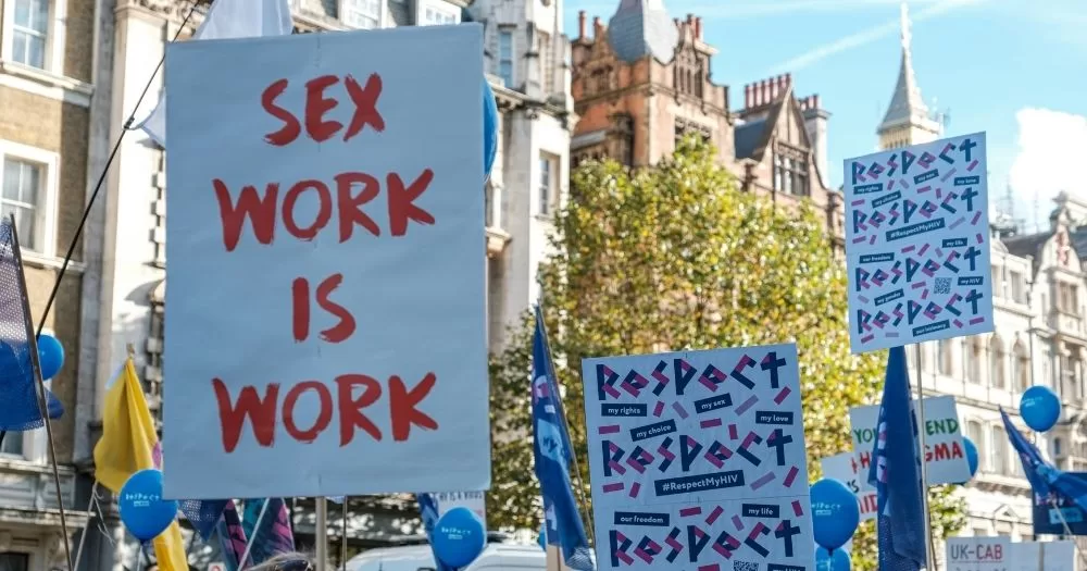 Image shows a protest to decimalise sex work. The camera is focused on a white sign with red writing that says 