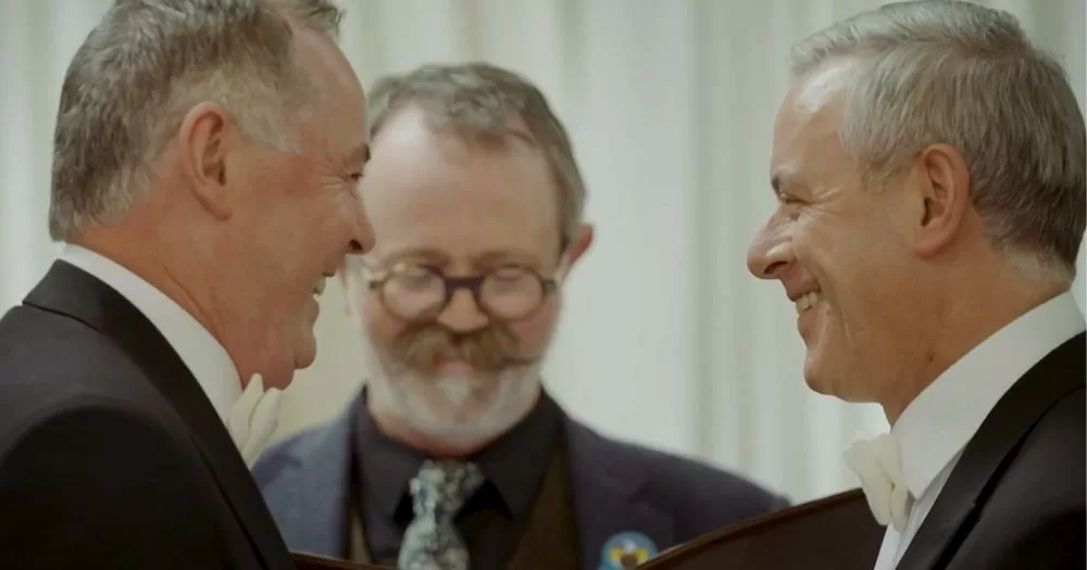 A screengrab from Glacaim Leat, the new Irish language documentary on MarRef. The image shows two smiley middle-aged men standing face to face at their wedding ceremony. Another stylish man stands between them, marrying the couple.