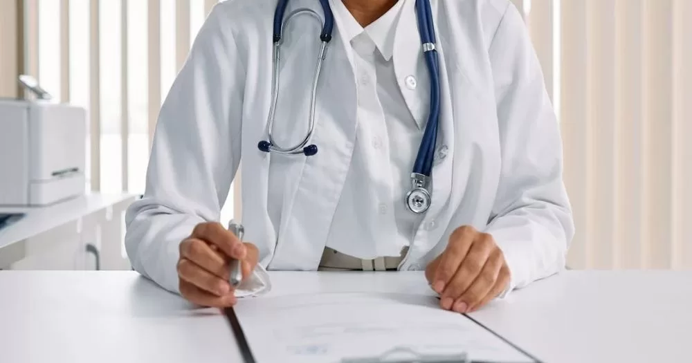This article is about healthcare inequalities in Ireland. In the photo, the arms and torso of a doctor writing on a clipboard while sitting at a desk.