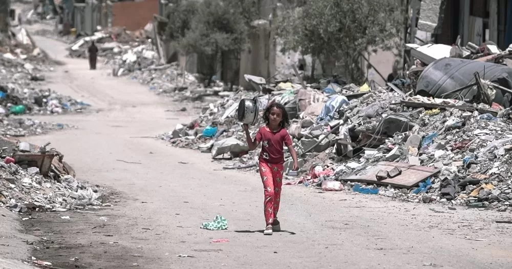 A child holds a pot and walks to an aid point in Gaza, surrounded by destruction.