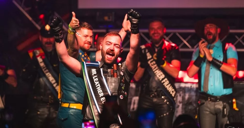 Image shows Jamie Ryan on stage at Mr Gay Europe wearing a sash that says Mr Gay Europe. He is surrounded by other competitors. Everyone is dressed in leather.
