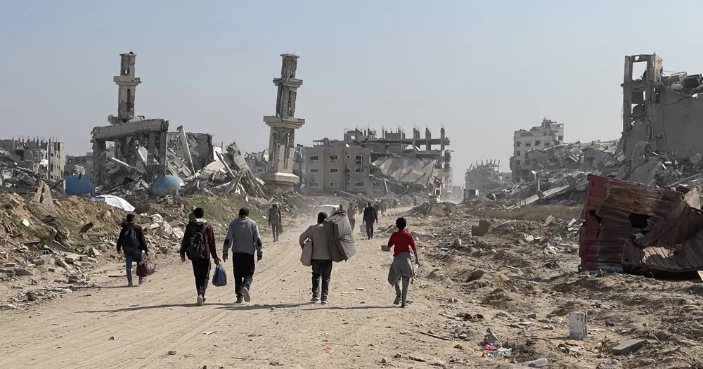 This article is about a Gaza ceasefire deal agreed upon by Israel and Hamas. In the photo, people in Gaza walking in a street with destroyed buildings in the background.