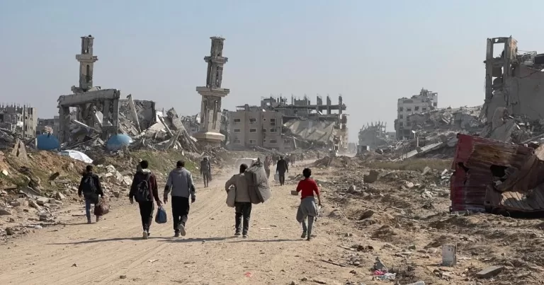 Image shows a group of displaced Palestinian people in Gaza carrying their belongings and walking surrounded by destruction caused by Israeli strikes.