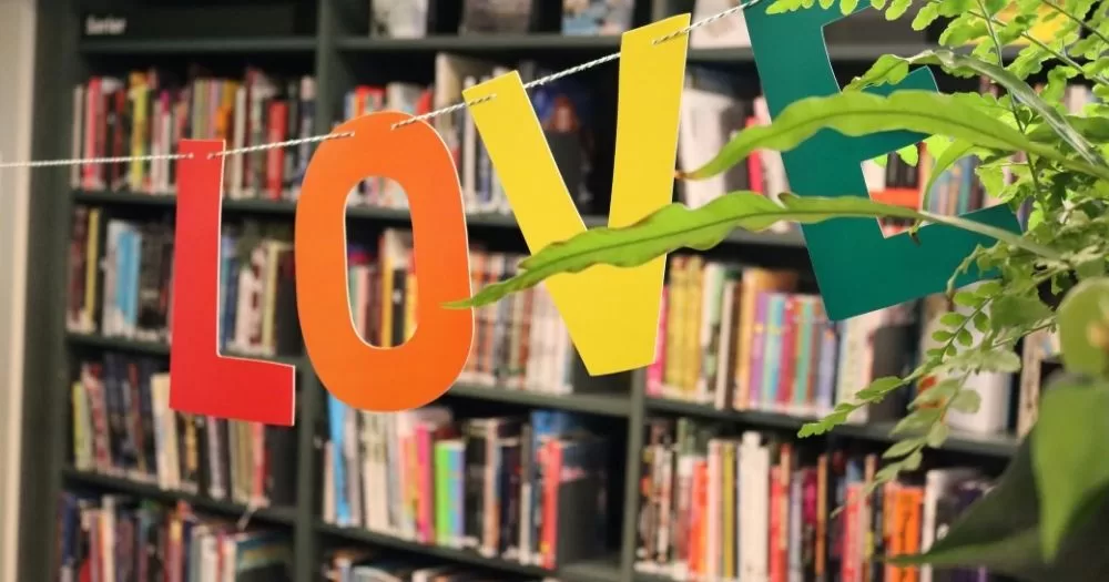 The image shows bunting saying 'Love', with a different colour for each letter, to represent the new LGBTQ+ library network. In the background are library shelves stocked with books. A plant can be seen in the right corner of the image.