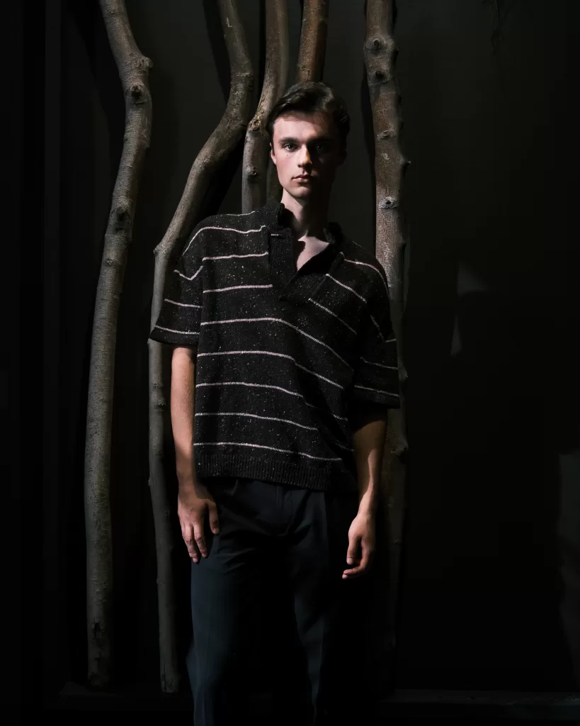 A model for Mar Knitwear's new collection with Donegal Yarns wears a sparkly black and gold striped polo style tee. They stand on front of trees on a black background.