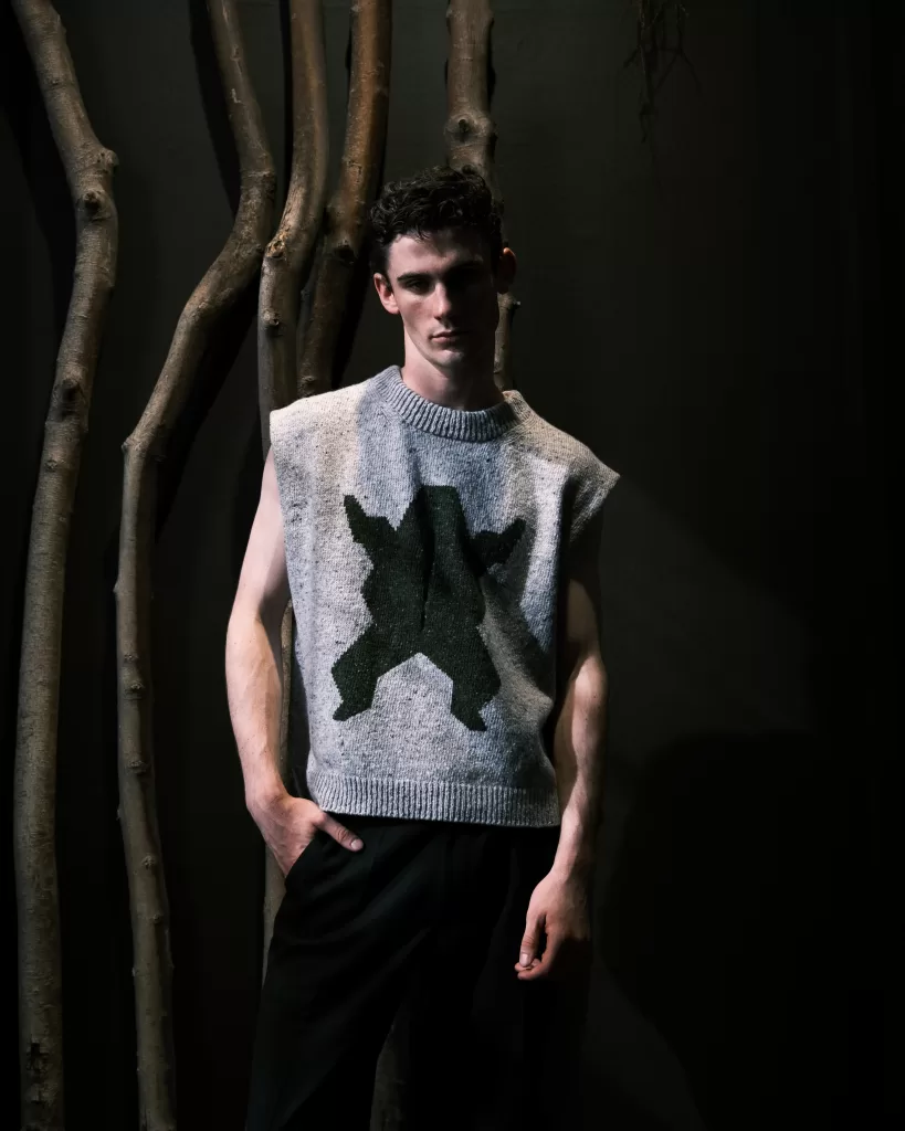 A model for Mar Knitwear's new collection with Donegal Yarns wears a grey vest with a frog on it. They stand on front of trees on a black background.