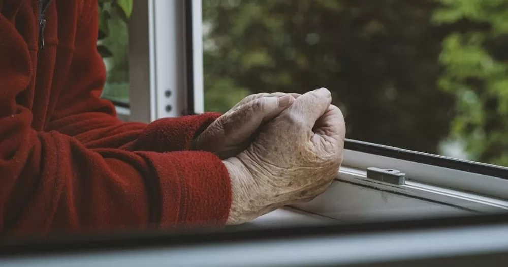 This article is about the need for reforms to protect LGBTQ+ older people in Ireland's care system. In the photo, the hands of an elderly person leaning on an open window while wearing a red top.