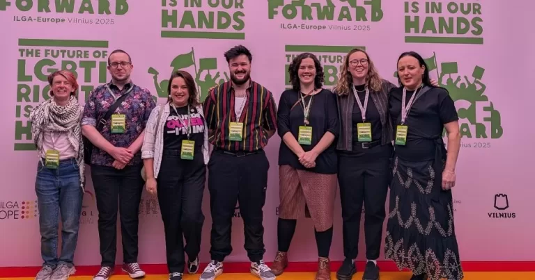 Image shows the Irish delegation at IGLA-Europe 2025 where it was announced that Outhouse will host next year's conference. They stand on a red carpet with a pink and green background.