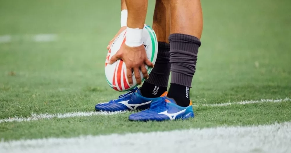 This article is about the Pride Round. In the photo, the feet and hands of a rugby player holding a rugby ball on the pitch.