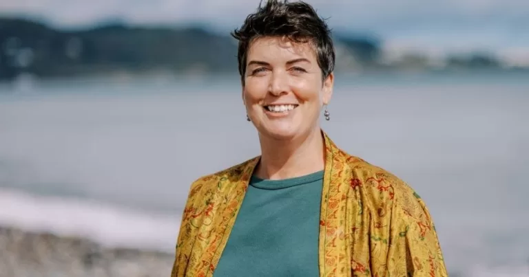 The image shows Ciara Foskin, author of poetry collection 'Meet the Follicles'. Her upper body is in frame, and she is wearing a blue shirt and yellow, silk jacket, smiling at the camera. She is standing on a beach, which is visible in the background.