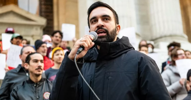 Zohran Mamdani holds a microphone and addresses a crowd at an anti-fascist rally. This image is being used to accompany an article about the mayor-elect's track record on LGBTQ+ rights.