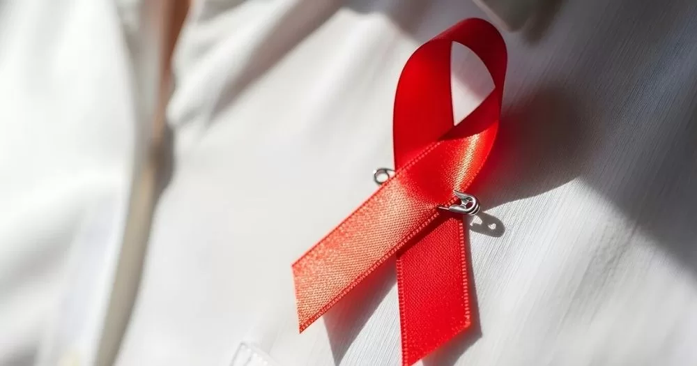 The image shows a red ribbon pinned onto a white shirt, with light shining on the ribbon creating pockets of shadow. The image is chosen to represent HIV, given the article's discussion of diagnoses in Ireland.
