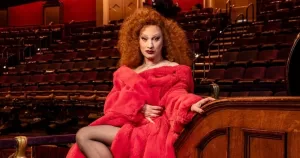 Jinkx Monsoon poses in an old theatre filled with red-upholstered seats. She wears a flowing red gown and heels. Her red hair is full of secrets.